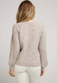 Pull en tricot beige clair avec une texture douce et duveteuse, manches longues et poignets côtelés. Design ajusté avec un ourlet arrière arrondi.