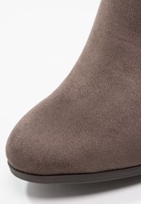 Gros plan sur l'avant d'une botte en daim taupe avec une forme arrondie et lisse et une semelle en caoutchouc foncé sur fond blanc.