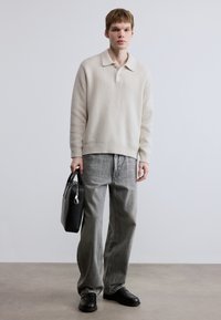 Ελαφρύ μπεζ πλεκτό πουλόβερ με γιακά, συνδυασμένο με γκρι τζιν φαρδιάς γραμμής. Το μοντέλο κρατάει μια μαύρη δερμάτινη τσάντα και φοράει σκούρα παπούτσια.