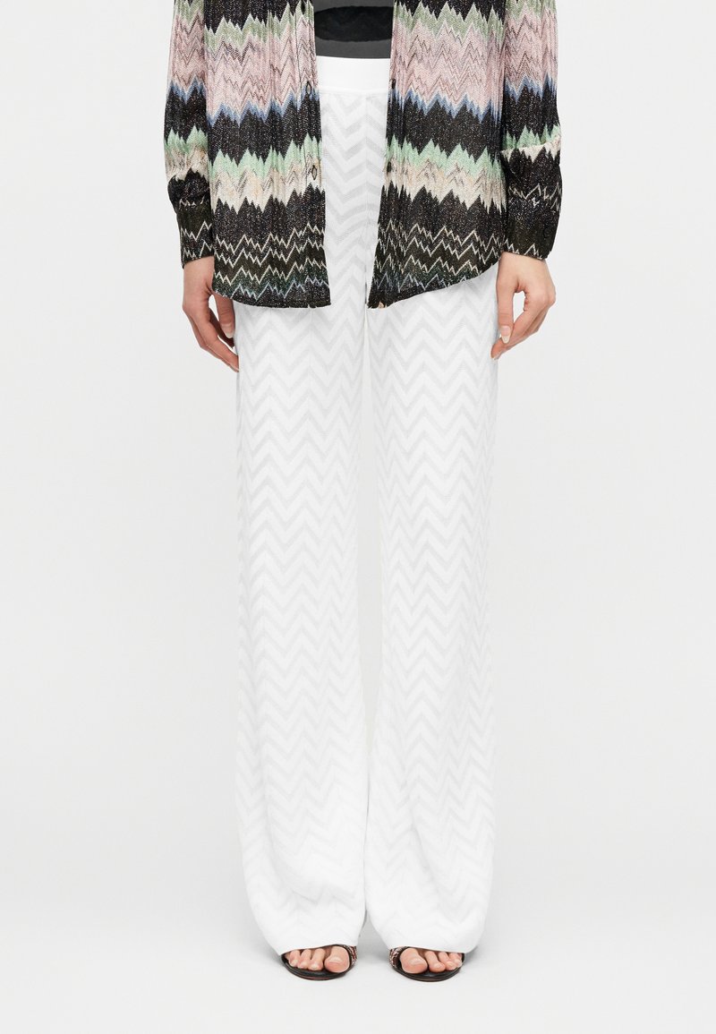 Γυναίκα που φοράει άσπρο παντελόνι φαρδιάς γραμ μής με υφή σε σχήμα ζιγκ-ζαγκ, πολύχρωμο ζακάρ ζακέτα με μοτίβο ζιγκ-ζαγκ και πέδιλα ανοιχτά στο πόδι, στέκεται μπροστά σε λευκό φόντο.