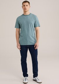 Kortærmet teal t-shirt med struktureret mønster, rund halsudskæring, kombineret med mørke jeans og sort-hvide sneakers.