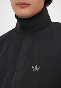 Crna jakna na zip s visokim ovratom, napravljena od glatkog materijala, s malim sivim logotipom na prsima. Vidljive su pojedinosti teksturirane površine.