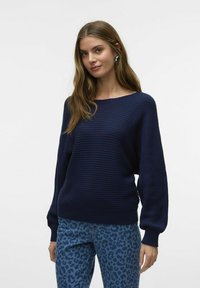 Pull en tricot côtelé bleu marine avec un large décolleté et des manches ballon, associé à un jean à imprimé léopard bleu clair. Texture lisse.