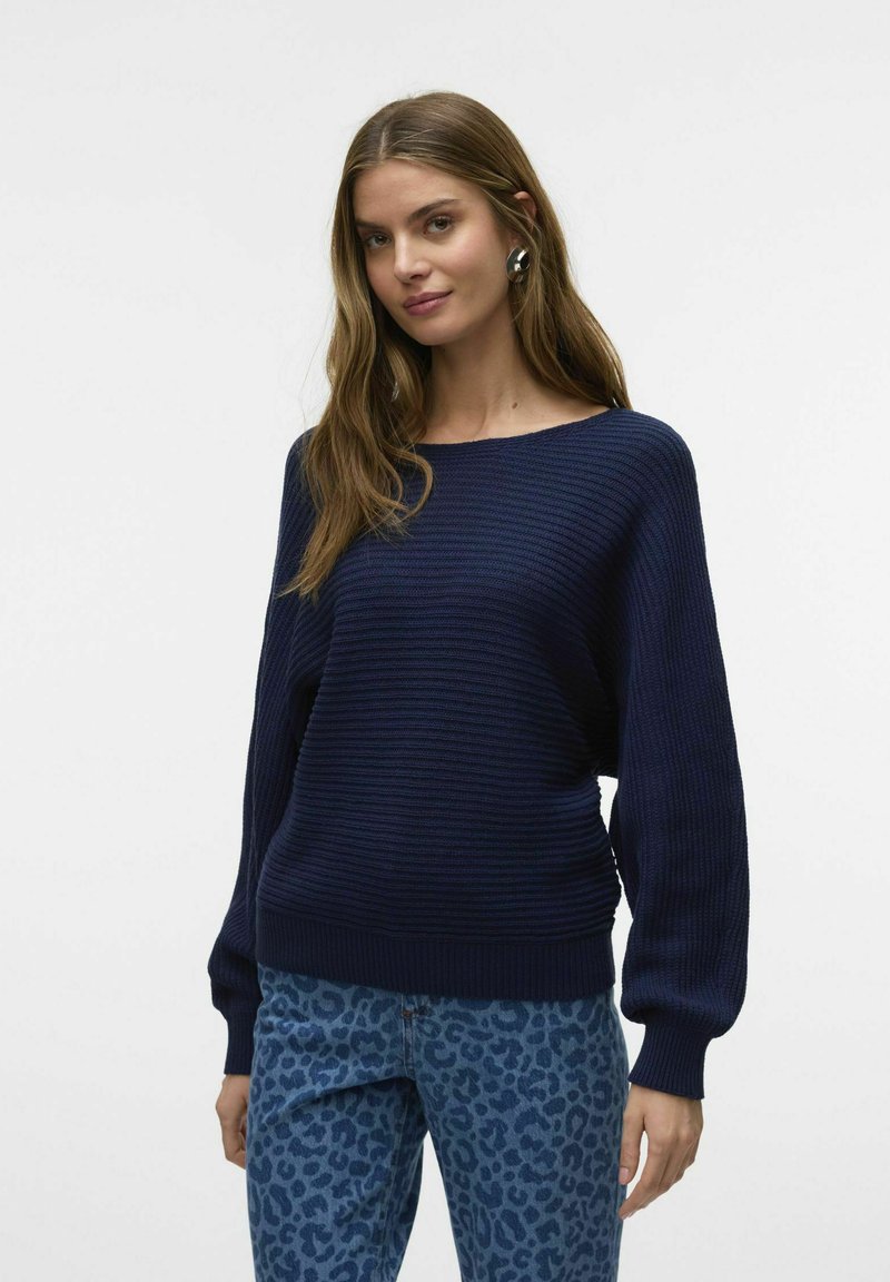 Pull en tricot côtelé bleu marine avec un large décolleté et des manches ballon, associé à un jean à imprimé léopard bleu clair. Texture lisse.