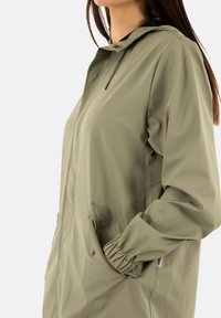 Chaqueta impermeable verde oliva con capucha, puños elásticos y bolsillos laterales. Textura suave con un acabado ligeramente brillante y detalles de cordón.