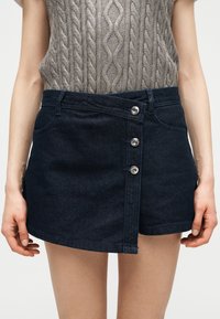 Γυναίκα που φοράει σκούρα τζιν μίνι φούστα με ασύμμετρο κούμπωμα με κουμπιά και μπροστινές τσέπες, συνδυασμένη με γκρι αμάνικο πλεκτό τοπ με σχέδιο καλώδιου.