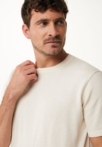 Krémové krátké tričko s kulatým výstřihem, které má jemnou texturu a volný střih, lehce držené rukou u výstřihu.