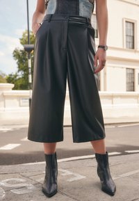 Crne culottes od umjetne kože s naboranim prednjim dijelom, širokim nogavicama i džepom. Uparene s teksturiranim crnim gležnjačama koje imaju šiljat vrh.