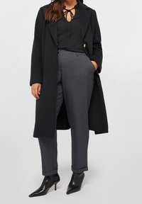 Musta pitkä takki mustan puseron päällä, jossa on solmukaulus, yhdistettynä harmaisiin räätälöityihin housuihin ja mustiin korkokenkiin, joissa on terävä kärki.