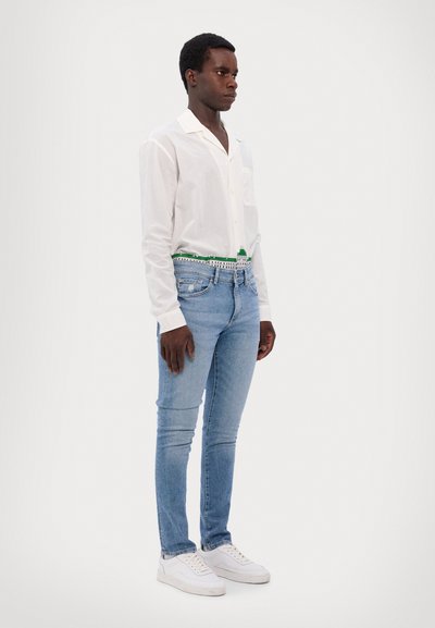 Bílá košile s dlouhým rukávem, světle modré skinny džíny, zelený vzorovaný opasek a bílé tenisky. Jednoduchý design, hladká textura, obepnutý střih.