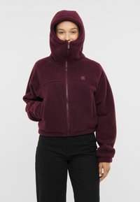 Burgundin värinen fleece-takki, jossa on korkea kaulus ja huppu, täydellinen vetoketju sekä etutaskut. Pehmeä pinta ja rento leikkaus.