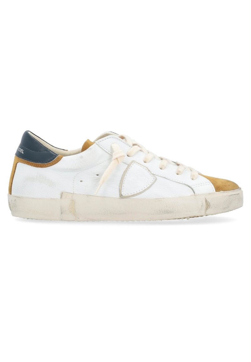 Sneaker en cuir blanc avec un embout beige, des accents caramel et un talon bleu marine. Dispose de lacets plats et d'une semelle en caoutchouc avec un aspect usé.