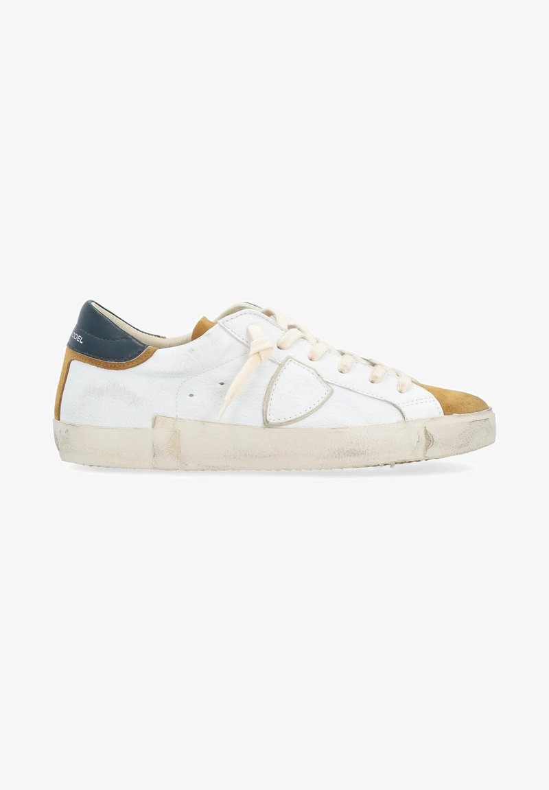 Sneaker en cuir blanc avec un embout beige, des accents caramel et un talon bleu marine. Dispose de lacets plats et d'une semelle en caoutchouc avec un aspect usé.