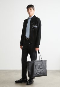 Svart ribbet cardigan med brodert logo, lyseblå skjorte, svarte bukser, skinnsko og en stor svart tote-bag med preget "HUGO."