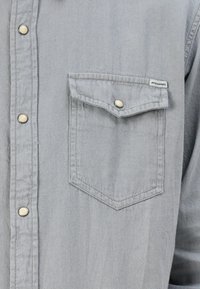 Camicia grigia con bottoni, dotata di tasca sul petto con patta, bottoni a pressione argentati e un tessuto strutturato. Etichetta visibile sulla tasca.