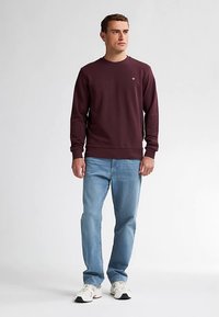 Viininpunainen pitkähihaisen ja pyöreäkauluksinen collegepaita, yhdistetty vaaleansinisiin farkkuihin ja valkoisiin tennareihin, päällä seisova mieshahmo.