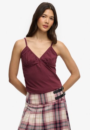 Jonge vrouw met lang donker haar, gekleed in een mouwloos bordeauxrode top met V-hals en een geplooide geruite rok met zwarte gespdetails.