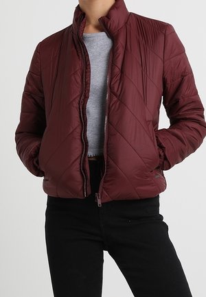 Osoba ubrana w bordową pikowaną kurtkę na szarym topie i czarnych spodniach, ręce w kieszeniach kurtki, stojąca na jednolitym tle.