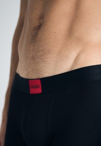 Schwarze Boxershorts mit einem schwarzen Bund, der ein rotes Patch mit dem Schriftzug "HUGO" trägt. Der Stoff ist glatt und enganliegend.