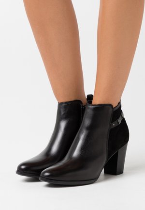 Botas de cuero negro hasta el tobillo con tacones cuadrados y detalle de hebilla, llevadas sobre piernas desnudas frente a un fondo blanco liso.