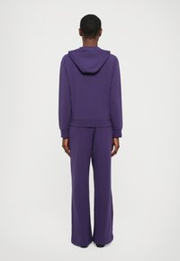 Paarse hoodie en bijpassende brede broek, beiden gemaakt van zachte stof, hebben ribgebreide boorden en een relaxed fit.