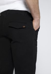 Pantaloni neri realizzati in un tessuto liscio, con una tasca posteriore dotata di una semplice patta e un piccolo accento arancione.