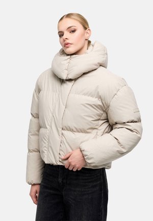 Donna con giacca imbottita beige con collo alto imbottito e pantaloni neri, in piedi su sfondo bianco.