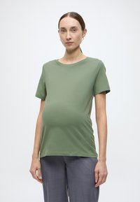 Μαλακό πράσινο μπλουζάκι εγκυμοσύνης με κοντά μανίκια, στρογγυλή λαιμόκοψη, εφαρμοστό στο πάνω μέρος και χαλαρή εφαρμογή στην περιοχή της κοιλιάς, συνδυασμένο με γκρι παντελόνι.