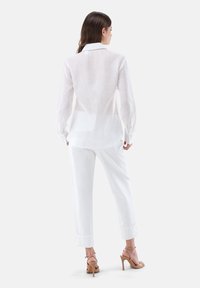 Camicia bianca in lino leggero con maniche lunghe, polsini abbottonati e un design con schiena aperta, abbinata a pantaloni larghi bianchi e sandali con tacco.