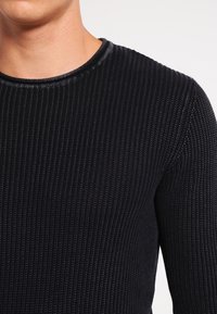 Camisa de punto negra con cuello redondo, que presenta una superficie texturizada y un diseño ajustado. La tela parece suave y elástica.