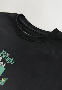 Schwarzes T-Shirt mit Rundhalsausschnitt und dem Grafikdesign "The Beagle", das eine Illustration eines Hundes und eines Vogels in gedämpften Grün- und Gelbtönen zeigt.