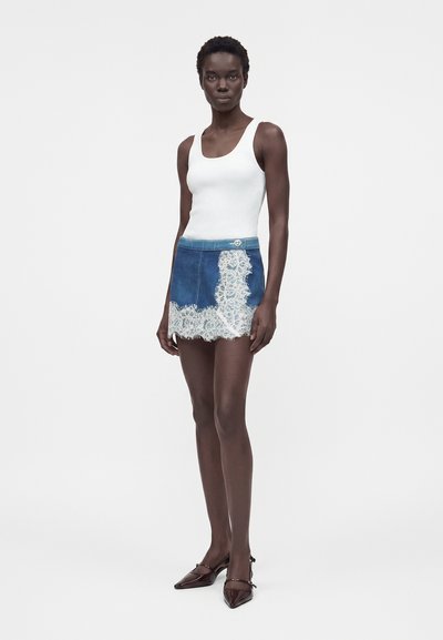 Γυναίκα που φοράει λευκό αμάνικο τοπ, μπλε μίνι τζιν φούστα με λευκή δαντέλα στη φάσα και σκούρα μυτερά τακούνια, στέκεται μπροστά σε μονόχρωμο φόντο.