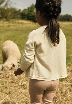 Jong kind met donker haar in een paardenstaart staat buiten naast een schaap dat uit een voederbak eet in een grasveld.