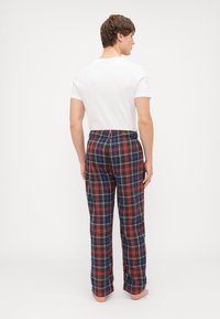 Un pantalon de pyjama à carreaux présente une base marine avec des lignes rouges, vertes et jaunes, une coupe décontractée et une ceinture élastique. Porté avec une chemise blanche.