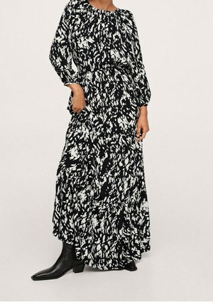 Frau, die ein langes schwarz-weiß gemustertes Kleid mit abstraktem Muster, langen Ärmeln und schwarzen Knöchelstiefeln trägt und mit einer Hand in der Tasche steht.