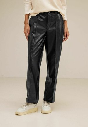 Osoba nosząca czarne, proste spodnie z imitacji skóry oraz białe trampki, stojąca na jednolitym beżowym tle.