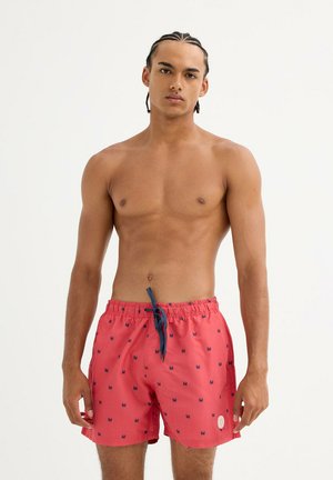 Joven con cabello trenzado que lleva pantalones cortos de baño rojos con pequeños patrones de cangrejos, de pie contra un fondo claro y liso.
