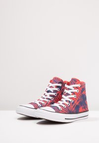 Zapatillas de caña alta de estampado tie-dye en rojo y azul marino con cordones blancos y suelas de goma sobre una superficie blanca contra un fondo liso.