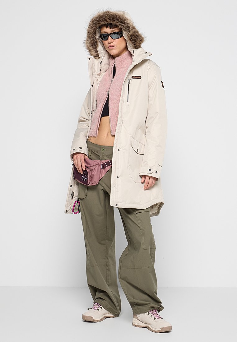 Beige wasserdichter Parka mit fellgefütterter Kapuze, über einem rosa cropped Fleece, olivfarbenen Cargo-Hosen und leichter Fußbekleidung. Kleine lila Taillen- Tasche.