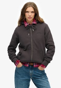 Sudadera con cremallera de color gris oscuro, con capucha ajustable, bolsillos frontales y logo. Llevada sobre una camisa de cuadros en tonos rosa y azul. Modelo con pantalones vaqueros ajustados de color azul.