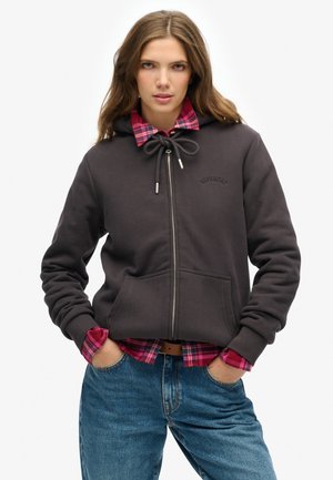 Dunkelgrauer Reißverschluss-Hoodie mit Kapuze und Kordelzug, Fronttaschen und Logo. Getragen über einem pink-blauen karierten Hemd. Modell in lässigen blauen Jeans.