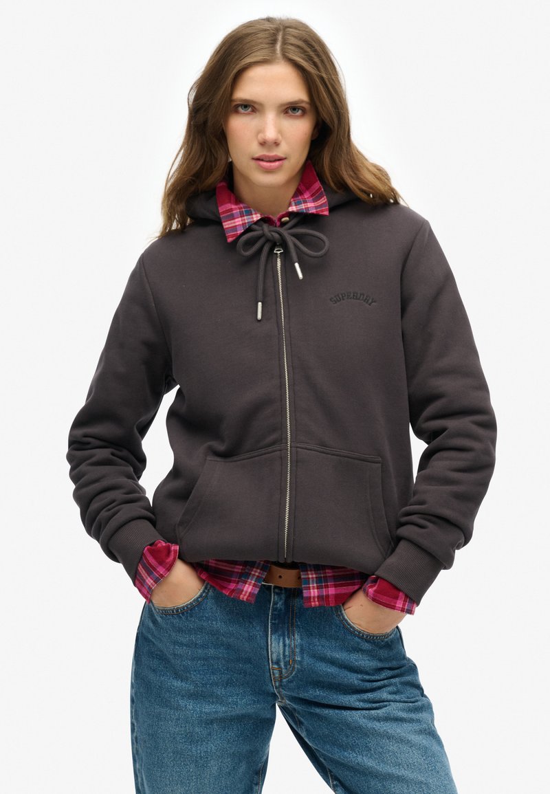 Sudadera con cremallera de color gris oscuro, con capucha ajustable, bolsillos frontales y logo. Llevada sobre una camisa de cuadros en tonos rosa y azul. Modelo con pantalones vaqueros ajustados de color azul.