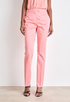 Kvinde iført pink bukser med højt talje og sidelommer samt sorte åbne sandaler mod en hvid baggrund.