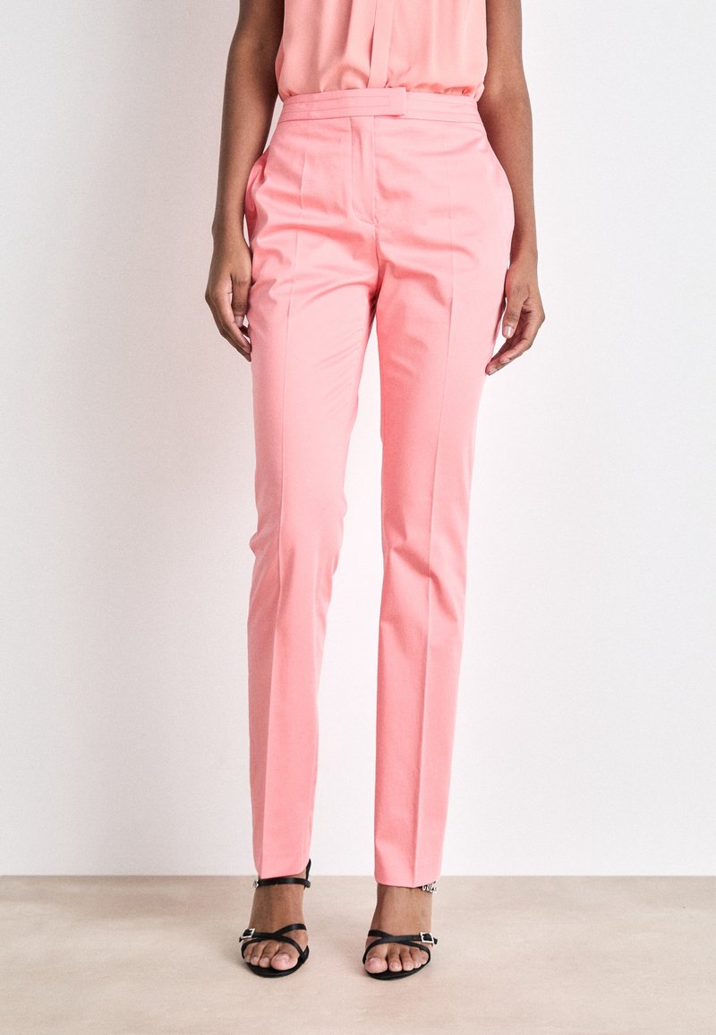 Femme portant un pantalon rose droit taille haute avec poches latérales et des sandales noires à bout ouvert sur fond blanc uni.