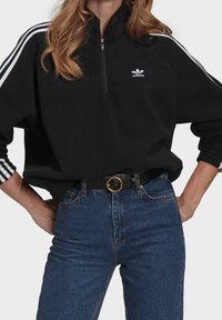 Musta fleece-collegepaita, jossa neljännekseen avautuva vetoketju, valkoinen Adidas-logo rinnassa ja valkoiset raidat hihoissa, yhdistetty korkeavyötäröisiin sinisiin farkkuihin ja mustaan vyöhön.