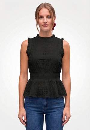 Frau mit blondem Haar, das mit einem roten Haargummi zurückgebunden ist, trägt ein ärmelloses schwarzes, strukturiertes Oberteil mit einem gerafften Saum und eine blaue Jeans vor weißem Hintergrund.