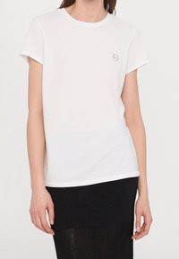 Bílá bavlněná tričko s krátkým rukávem, kulatým výstřihem a malým kulatým logo na levé straně hrudi. Hladká textura, jednoduchý design.