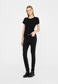 Donna in piedi che indossa un top nero decorato a maniche corte, pantaloni neri aderenti, stivaletti neri e orecchini pendenti argentati su sfondo bianco.
