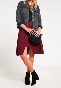 Veste en denim noire, haut noir ajusté, jupe bordeaux avec fente sur le côté, bottines noires, et sac à bandoulière rond noir.