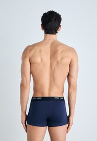 Marineblaue Herrenboxershorts aus glattem Stoff, mit einem schwarzen Bund, auf dem der Markenname "HUGO" deutlich sichtbar ist.